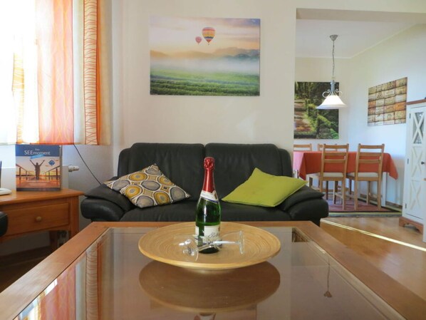 A living room with a glass coffee table, a bottle of champagne, a sofa with patterned cushions, and a painting of hot air balloons. - Holiday Apartment Seagull (Bad Saarow)