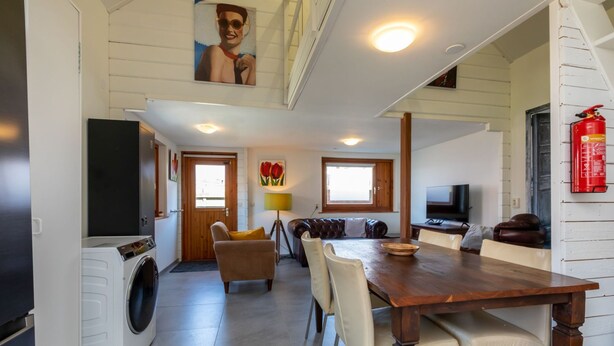 A compact kitchen with a washing machine, a dining area with a wooden table and chairs, and a TV mounted on the wall.