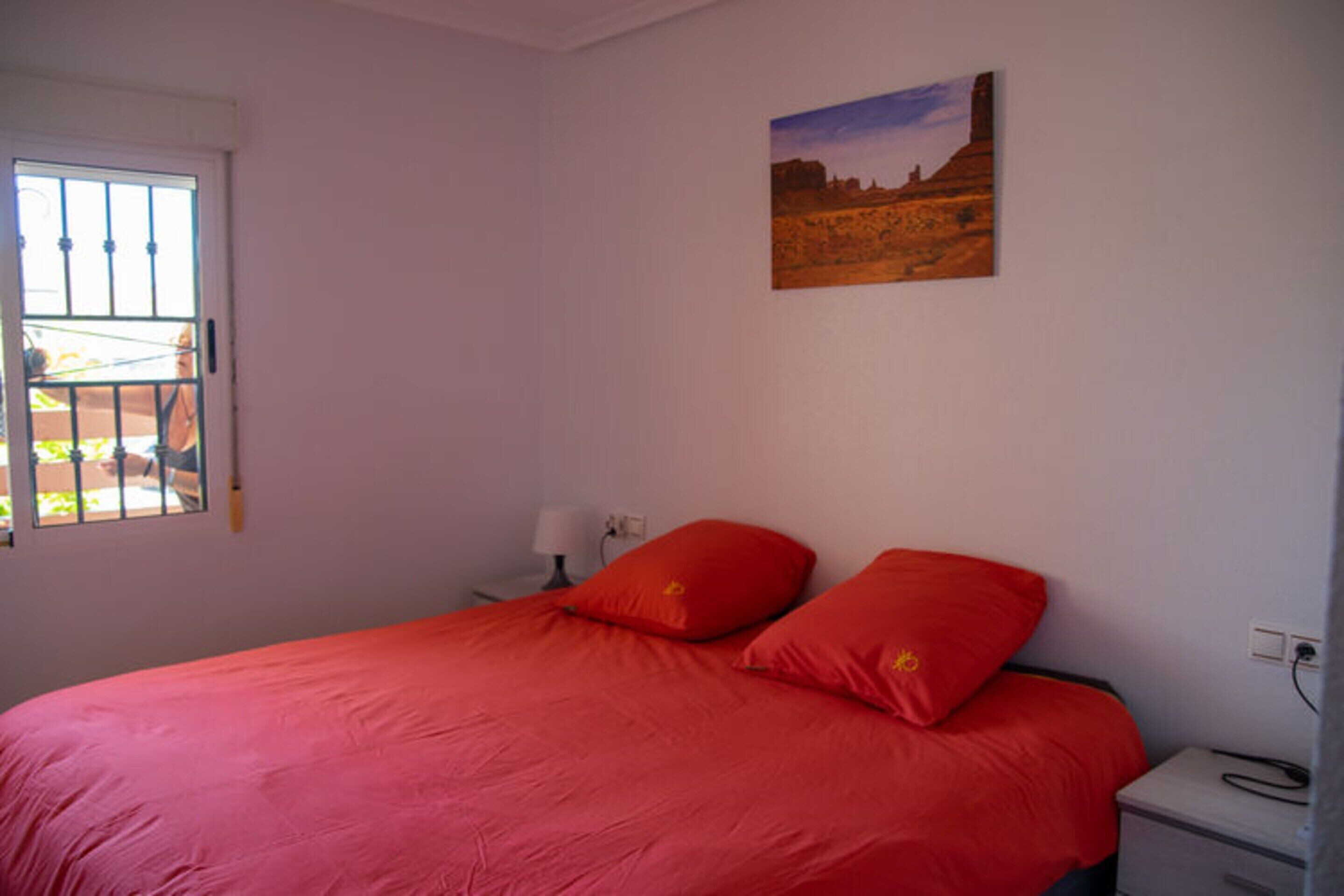 Un dormitorio con una cama, una mesita de noche, una lámpara, una ventana con rejas y un cuadro en la pared.