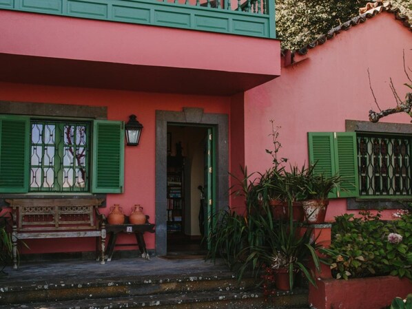 A building with a pink facade, green shutters, and a small porch with a bench and potted plants. - Country House in Gran Canaria With Private Pool (Vega de San Mateo)