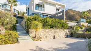 Una moderna casa a due piani con un vialetto in pietra che conduce all'ingresso, immersa nel verde e con le montagne sullo sfondo.