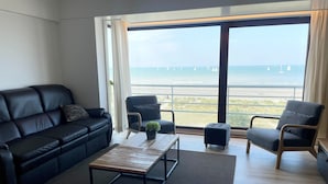 Uma sala de estar moderna com um sofá de couro preto, uma mesa de centro de madeira e duas poltronas. Uma grande janela oferece vista para a praia e barcos à vela.