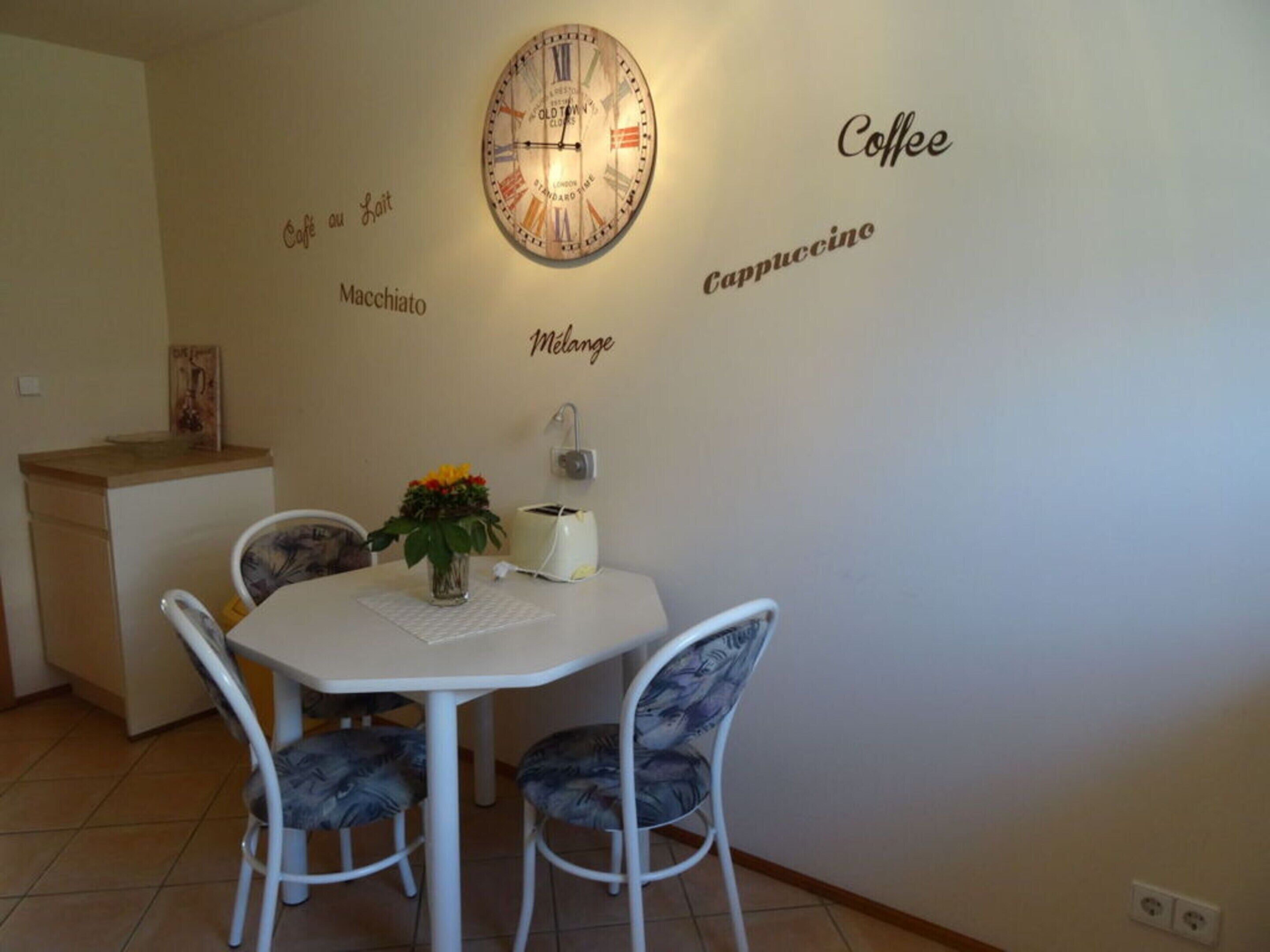 A dining area with a white table, two chairs, a clock, and wall decorations reading "Coffee," "Cappuccino," "Macchiato," and "Melange."