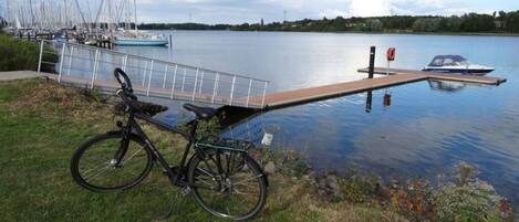 A bicycle with a cargo rack parked on a grassy area by a calm lake, with boats docked in the background.