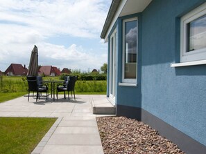A patio with a table and chairs, a small garden area with pebbles, and a blue building with windows. - Fereienhaus Mellumplate (Wurster Nordseeküste)