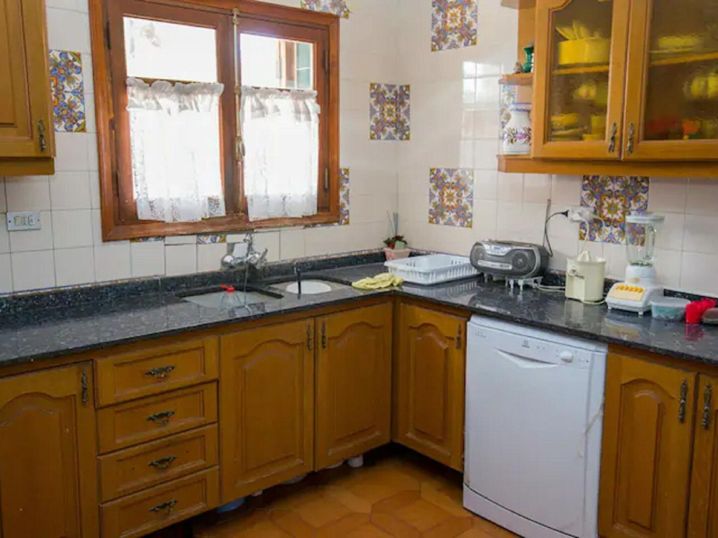 A kitchen with wooden cabinets, a white dishwasher, a sink, and a window with curtains.
