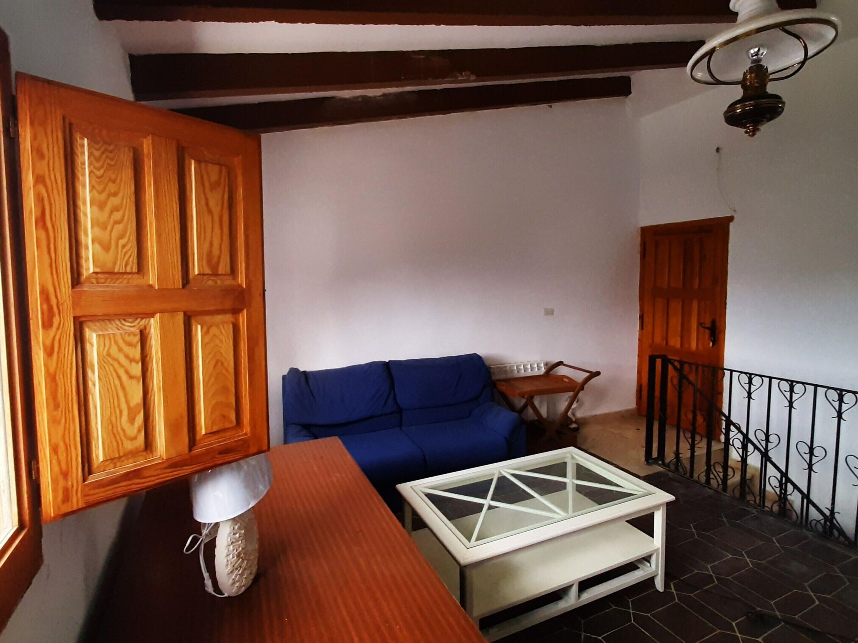 Una habitación con una puerta de madera, un sofá azul, una mesa blanca con superficie de vidrio y una escalera con barandilla negra.