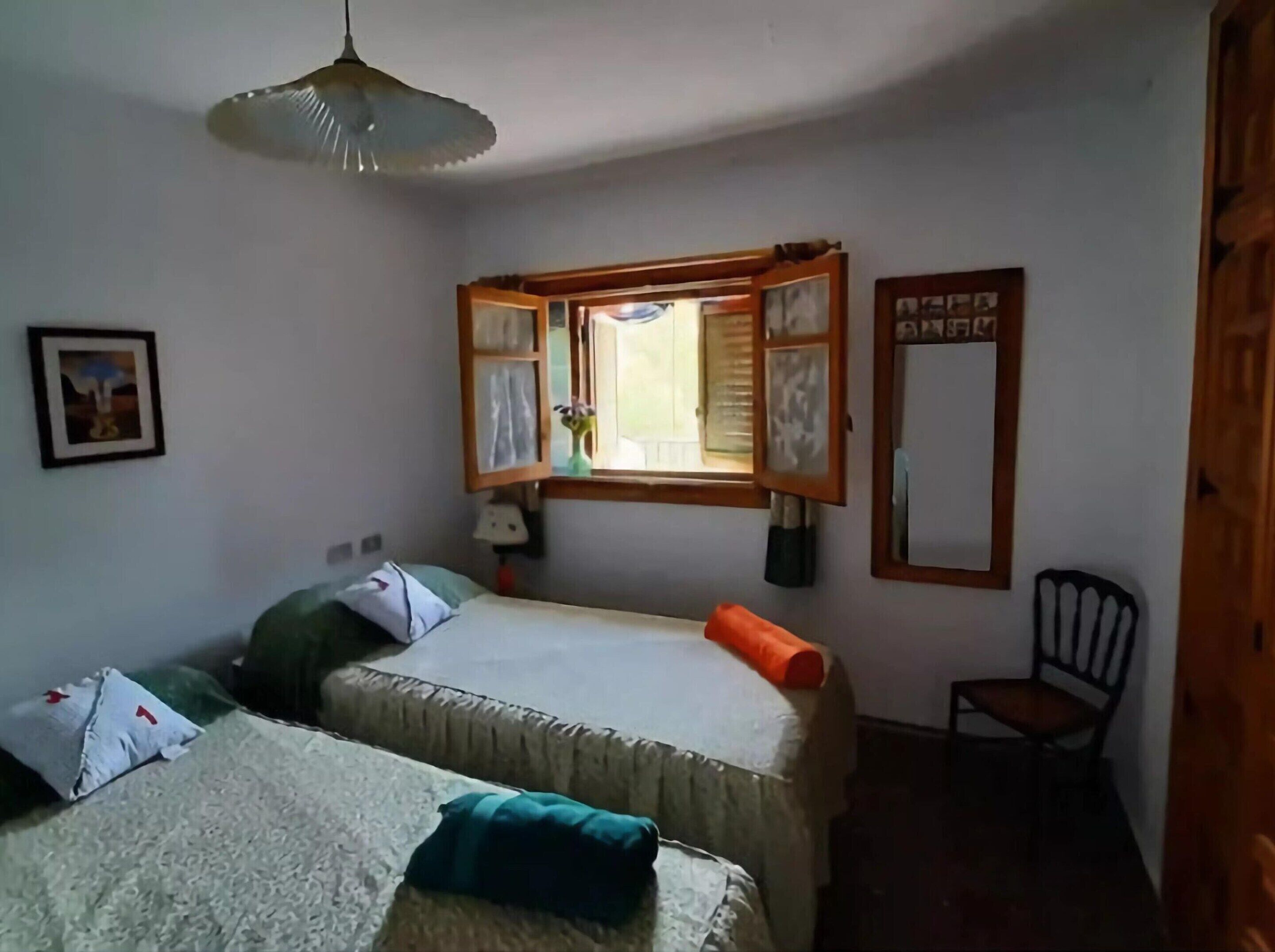 Una habitación con dos camas individuales, una ventana con marco de madera, una silla y una mesita.