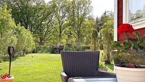 Eine Terrasse mit Korbmöbeln, einem Topf mit roten Blüten und Blick auf einen gepflegten Rasen und Bäume.