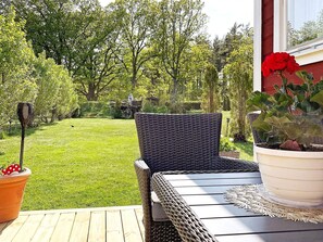 A patio with a wicker table and chairs, a potted plant with red flowers, and a view of a well-maintained lawn and trees. - 4 Person Holiday Home in Gusum (Valdemarsvik)