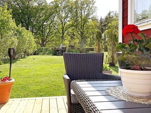 Une terrasse avec une table et des chaises en osier, un pot de fleurs aux fleurs rouges, et une pelouse avec des arbres et un chariot de jardin en arrière-plan.