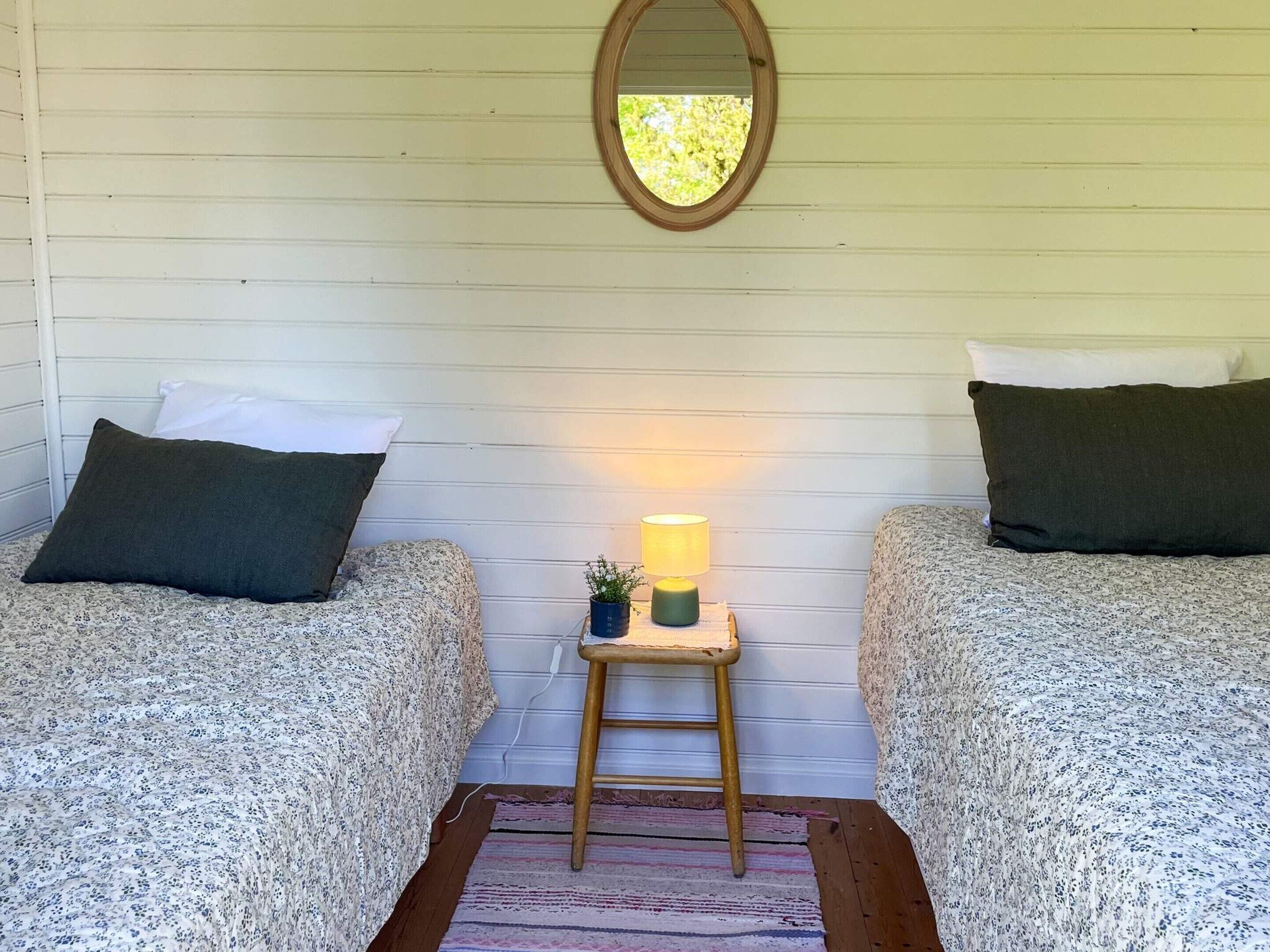 Una habitación con dos camas, una mesita pequeña con una lámpara, un espejo en la pared y suelo de madera.
