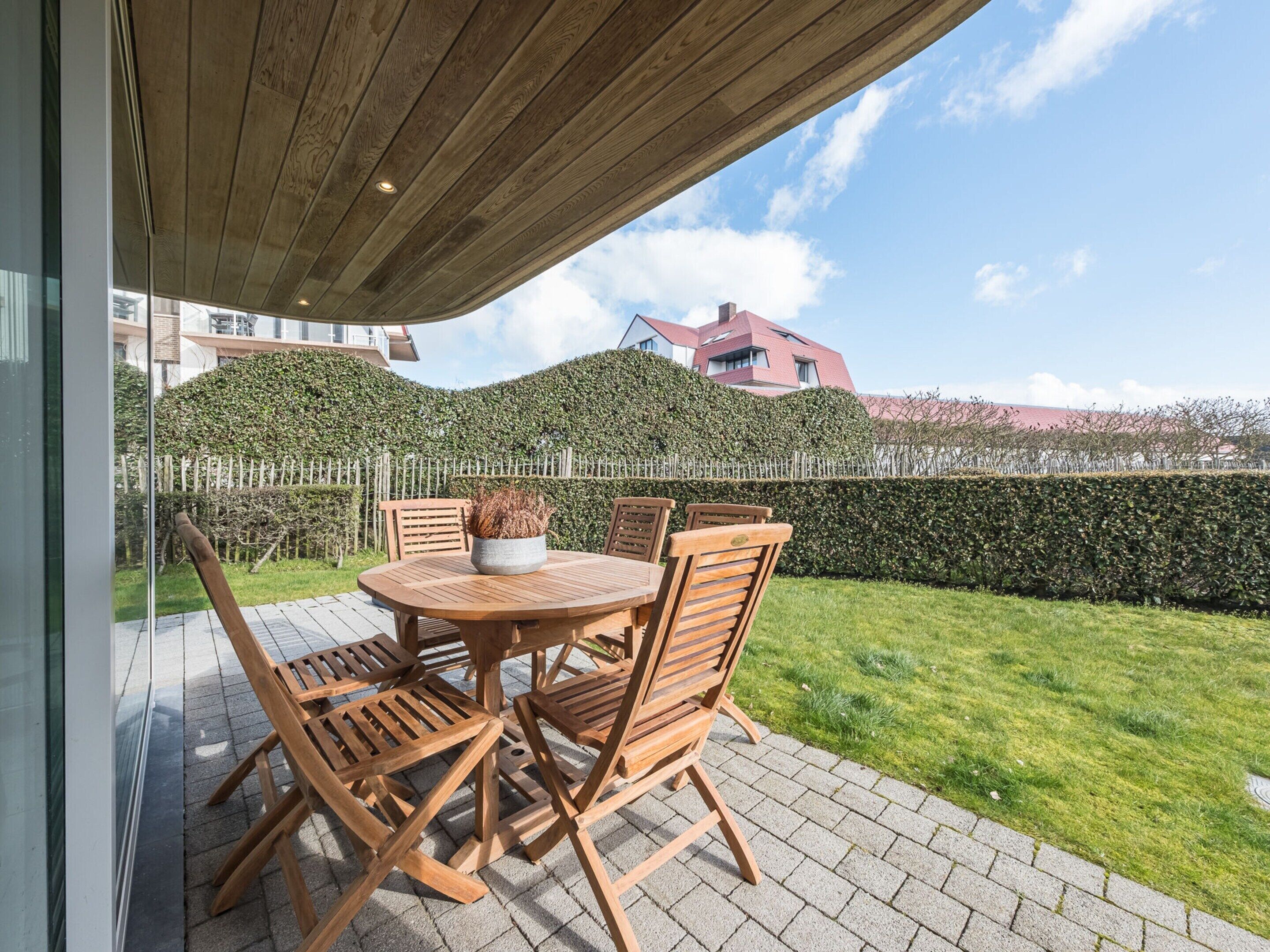 Une terrasse extérieure avec une table et des chaises en bois rondes, un pot de fleurs et une vue sur un arbuste taillé avec soin et des bâtiments en arrière-plan.