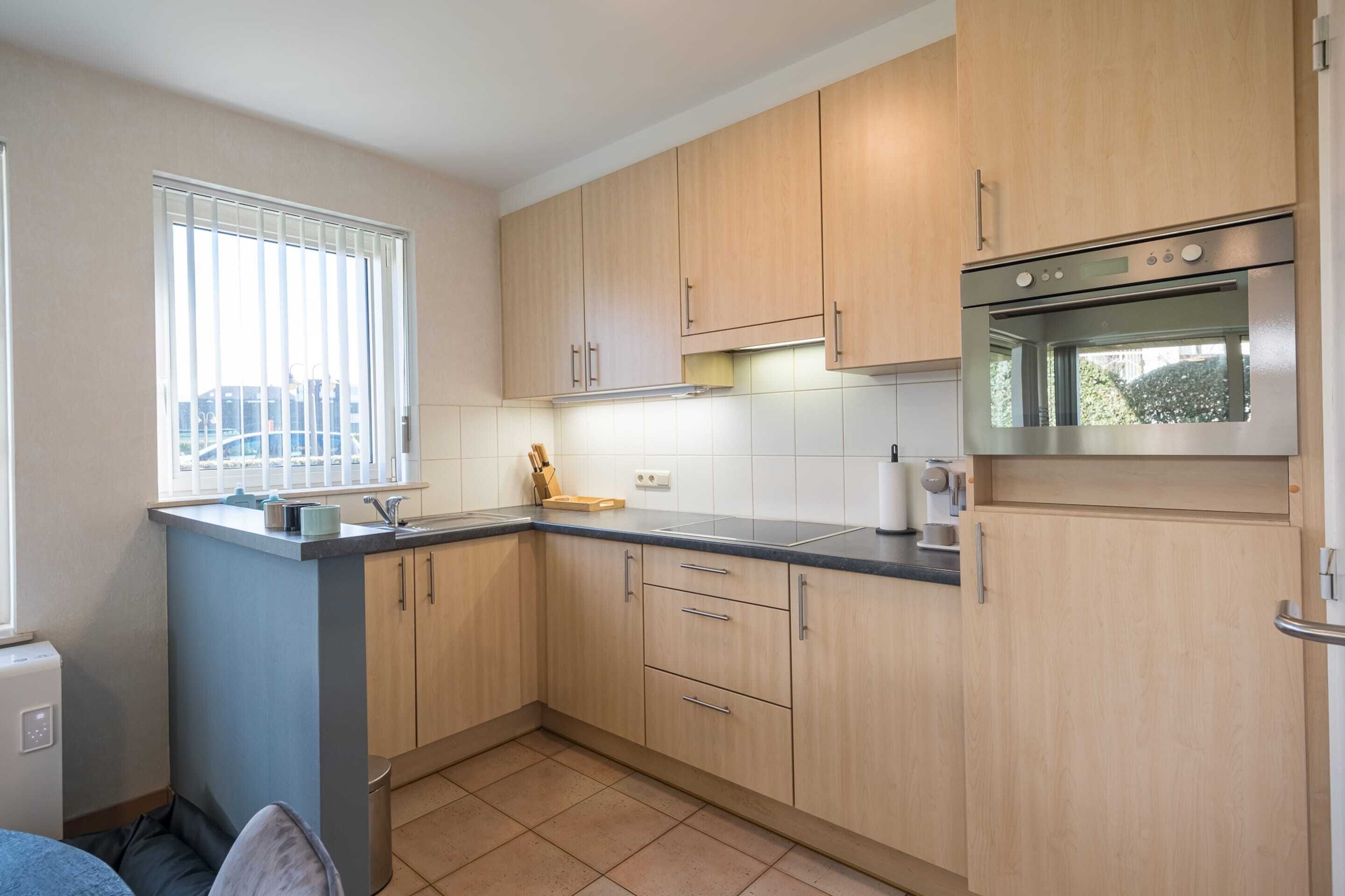 A compact kitchen with light wood cabinets, a stainless steel oven, and a window with blinds.