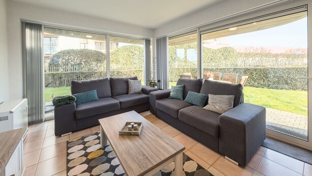 Un salon moderne avec un canapé d’angle, une table basse et de grandes fenêtres donnant sur un jardin.