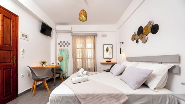 A hotel room with a bed, a desk, a chair, a television, and a window with curtains. - Marina's studios (Santorini)