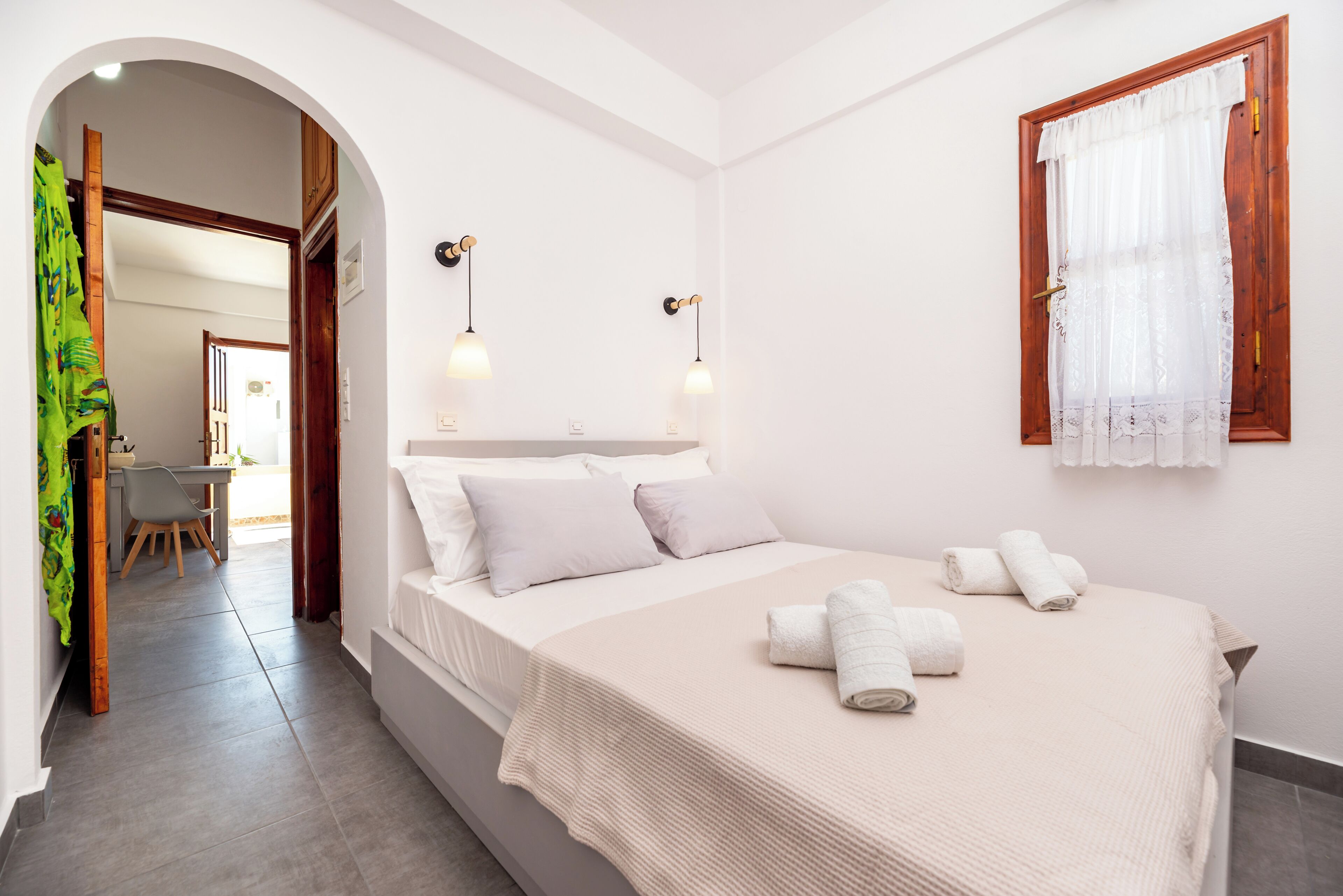 A bedroom with a bed, two bedside lamps, a window with a curtain, and a desk with a chair.
