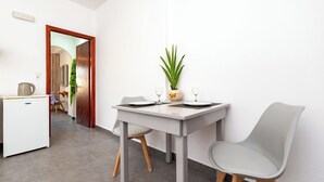 A modern dining area with a white table, two grey chairs, a potted plant, and a small kitchenette in the background. - Marina's studios (Santorini)
