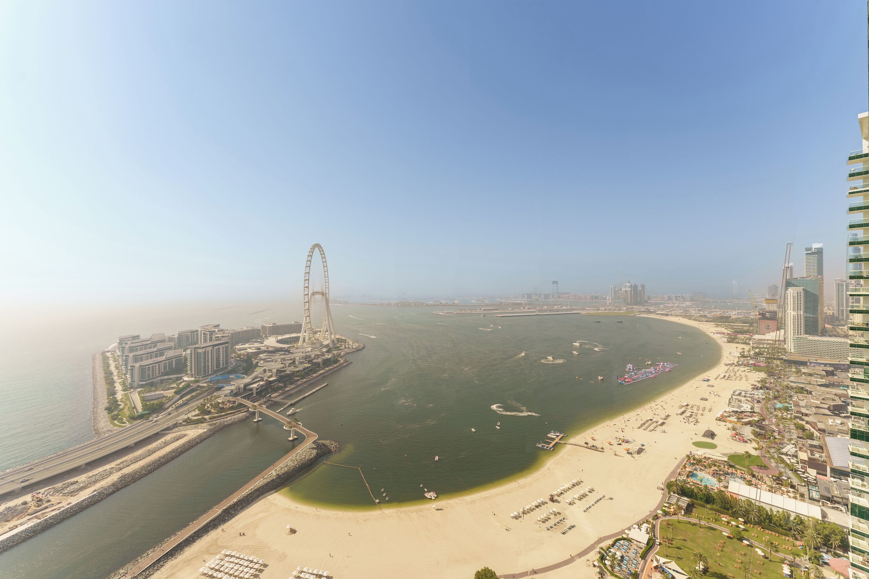 An aerial view of a coastal city with a prominent Ferris wheel, a beach, and modern high-rise buildings.