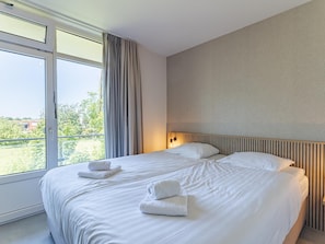 A hotel room with a large bed, a window with curtains, a wall-mounted light, and a view of greenery outside. - Zoutelande Retreat by Sea (Zoutelande)