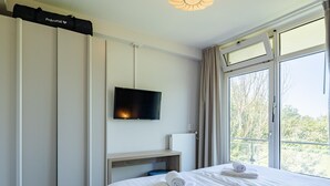A hotel room with a bed, a flat-screen TV mounted on the wall, a window with a view of trees, and a ceiling light fixture. - Zoutelande Retreat by Sea (Zoutelande)