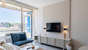 A modern living room with a sofa, a coffee table, a TV mounted on the wall, and a view of the outside. - Zoutelande Retreat by Sea (Zoutelande)