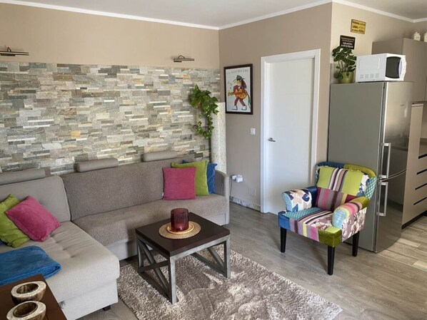 A living room with a stone wall, a sofa with colorful cushions, a small table, a colorful chair, and a refrigerator.