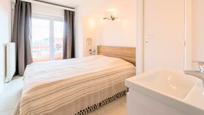 A bedroom with a bed, a nightstand, a wall-mounted lamp, a window with curtains, and a view of buildings outside. - Koksijde Beachfront Vitalo 502 (Koksijde)
