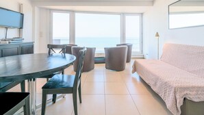 A modern living room with a dining area, large windows with a view of the sea, a flat-screen TV, and a sofa. - Koksijde Beachfront Vitalo 502 (Koksijde)