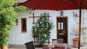 Une terrasse en bois avec un parasol rouge, une table ronde entourée de chaises, un pot de fleurs et un rosier grimpant.