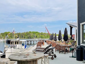 A waterfront patio with wooden tables and chairs, a boat docked at the pier, and a crane in the background.