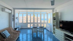 A spacious living room with a large window, a ceiling fan, a flat-screen TV, a dining area, and a sofa.