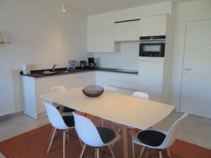 A modern kitchen with white cabinets, a black countertop, and a built-in oven. There is a dining table with four chairs and a decorative bowl on it. - Nieuwpoort Beach Getaway (Koksijde)