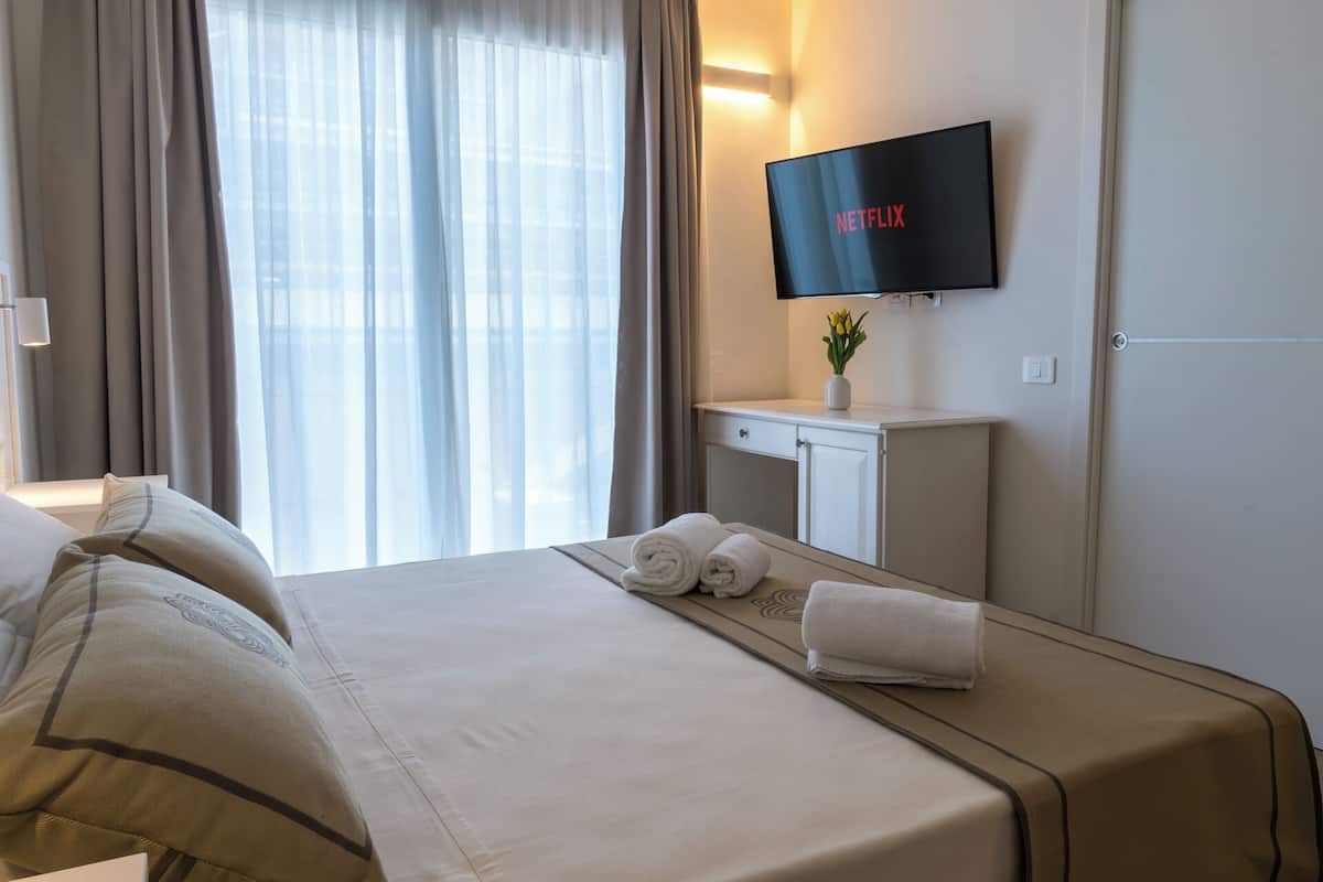 Ein Hotelzimmer mit Bett, einem an der Wand befestigten Fernseher, der 'NETFLIX' zeigt, einem kleinen Tisch mit Blumenvase und einem Fenster mit Vorhängen.
