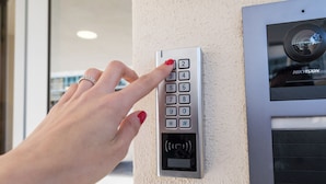 Una mano pulsando un teclado numérico en un dispositivo fijado a la pared, probablemente un cerrojo o interfón, con una cámara de seguridad HIKVISION encima.