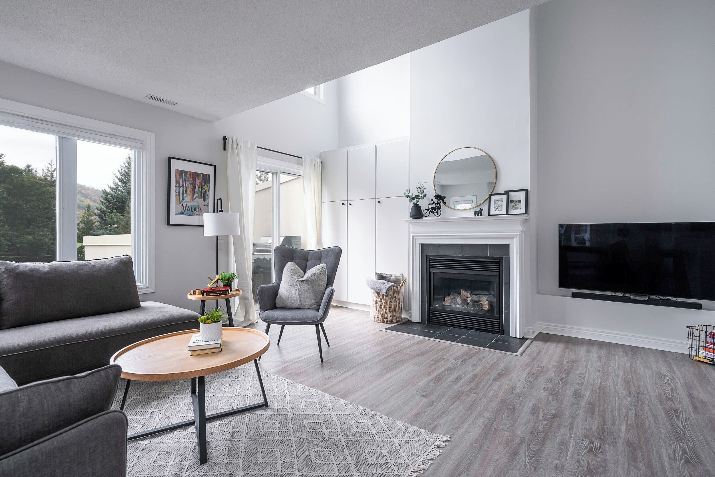 Una sala moderna con un sofá gris, una mesa de centro redonda de madera, un sillón gris y una chimenea con un televisor encima.