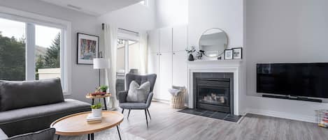 Una sala moderna con un sofá gris, una mesa de centro redonda de madera, un sillón gris y una chimenea con un televisor encima.