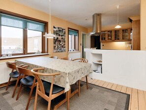 Un coin repas avec une table dressée pour quatre personnes, des chaises en bois, une nappe à motifs et un espace cuisine doté de meubles en bois et d’une hotte aspirante.