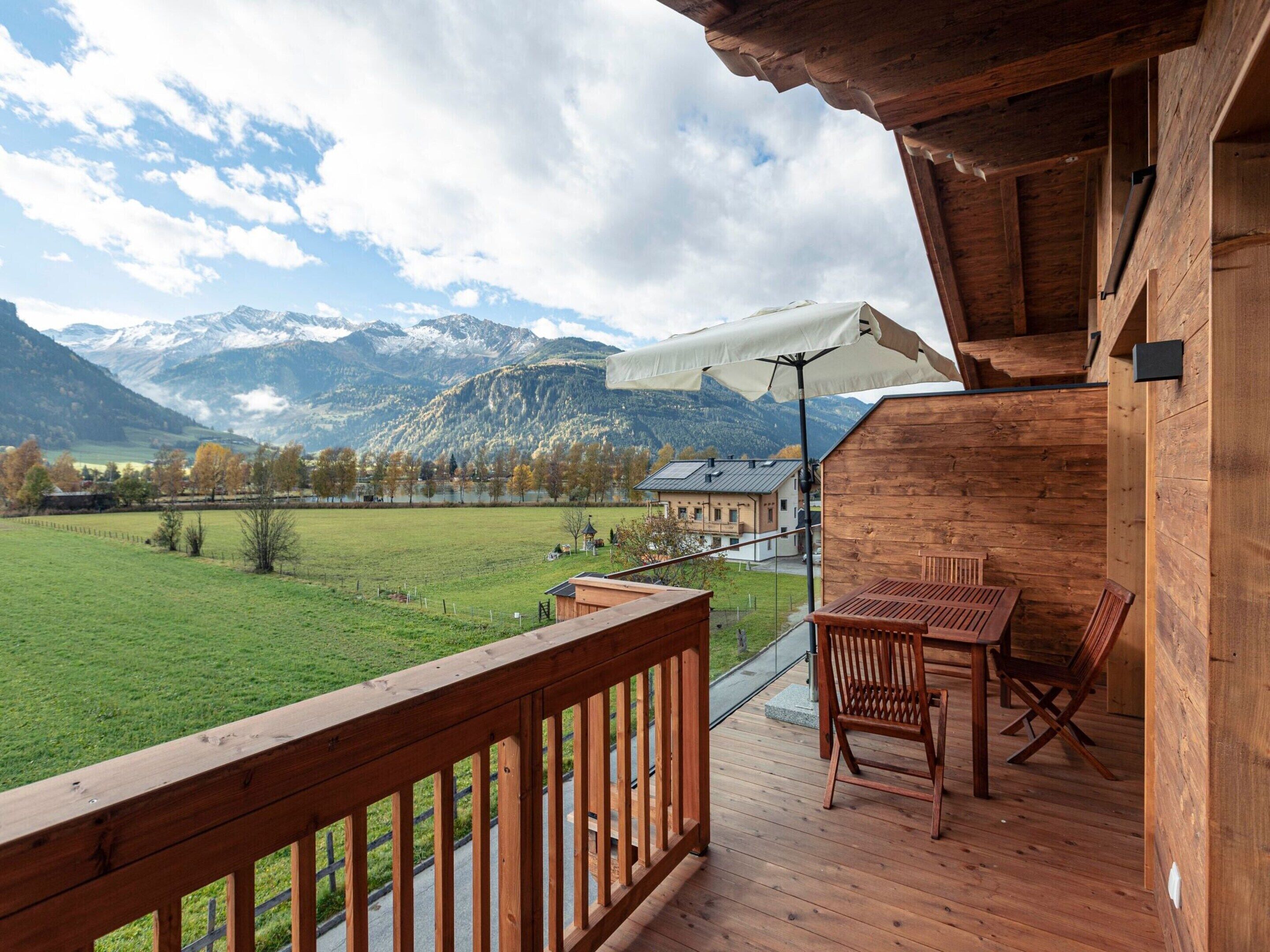Un terrazzo in legno con tavolo e sedie, affacciato su un prato verde e montagne.