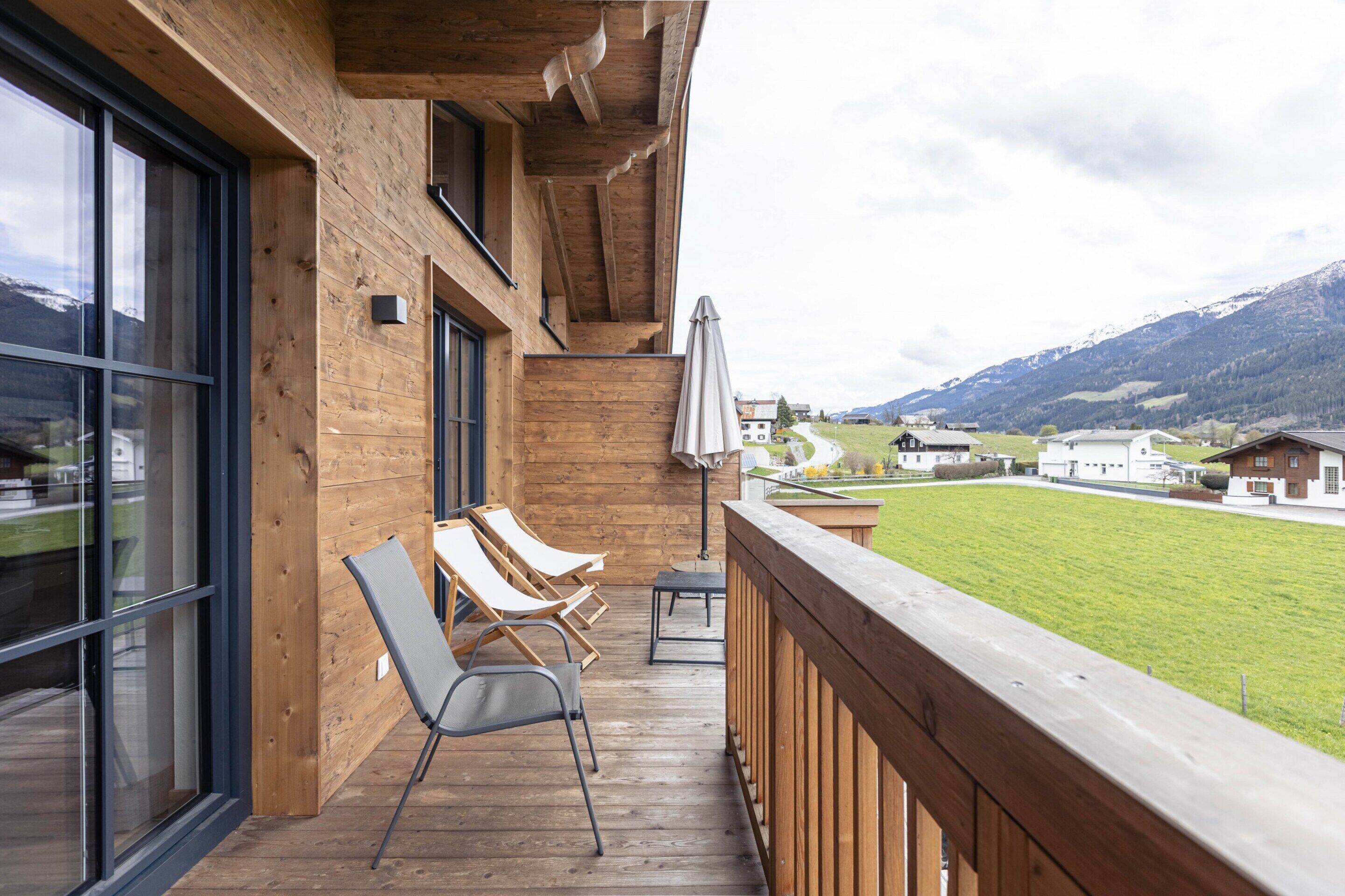 A wooden deck with lounge chairs and a table, overlooking a grassy area and mountains.