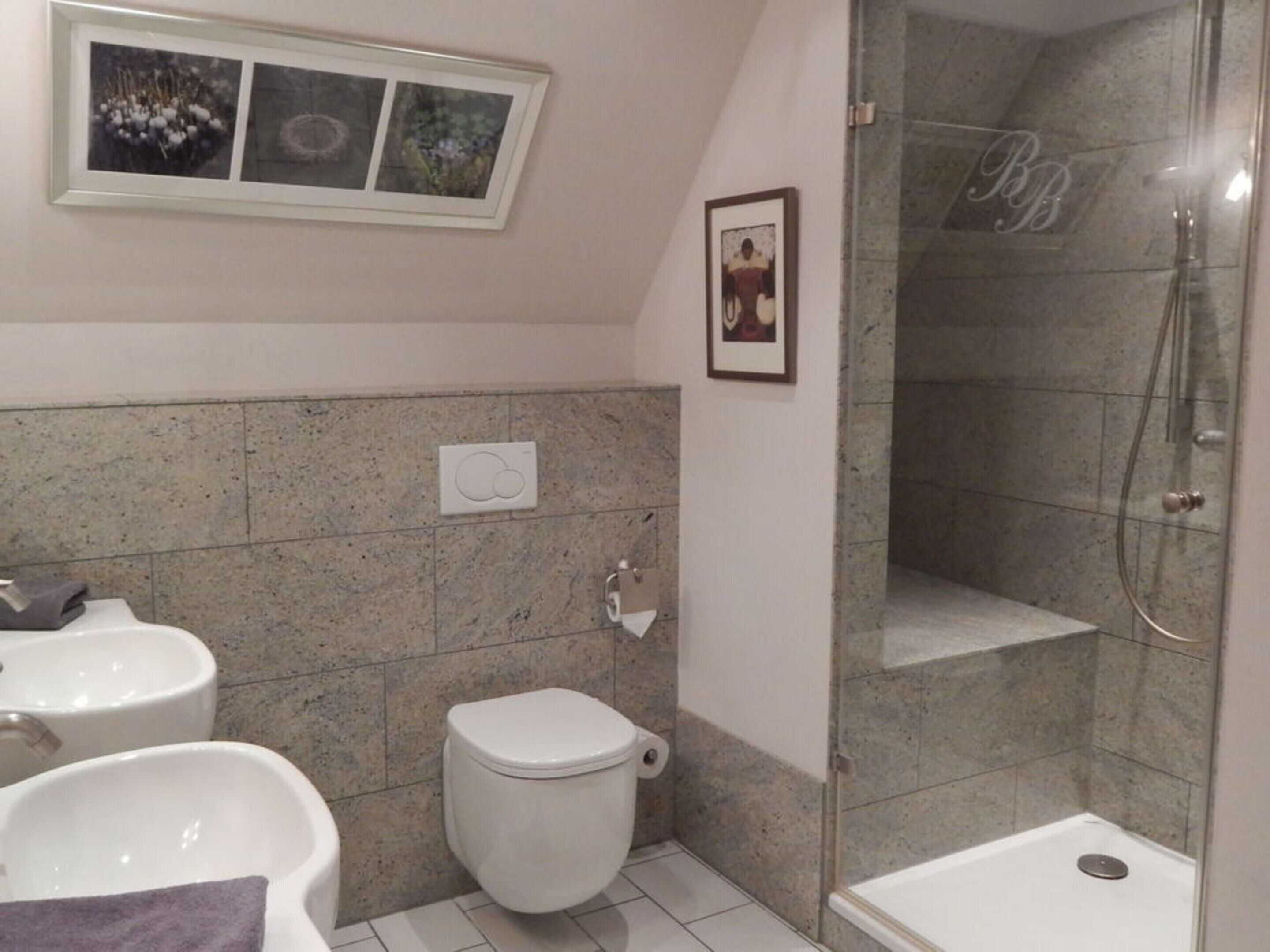 Une salle de bain équipée de toilettes, d’un bidet et d’une douche. Des tableaux encadrés ornent le mur.