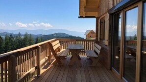 Eine Holzdielen-Terrasse mit Tisch und Bänken, mit Blick auf eine gebirgige Landschaft.