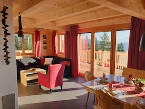 A cozy cabin interior with a wooden dining table set for two, a red armchair, and a sofa with red cushions. Large windows offer a view of a wooden deck and trees.