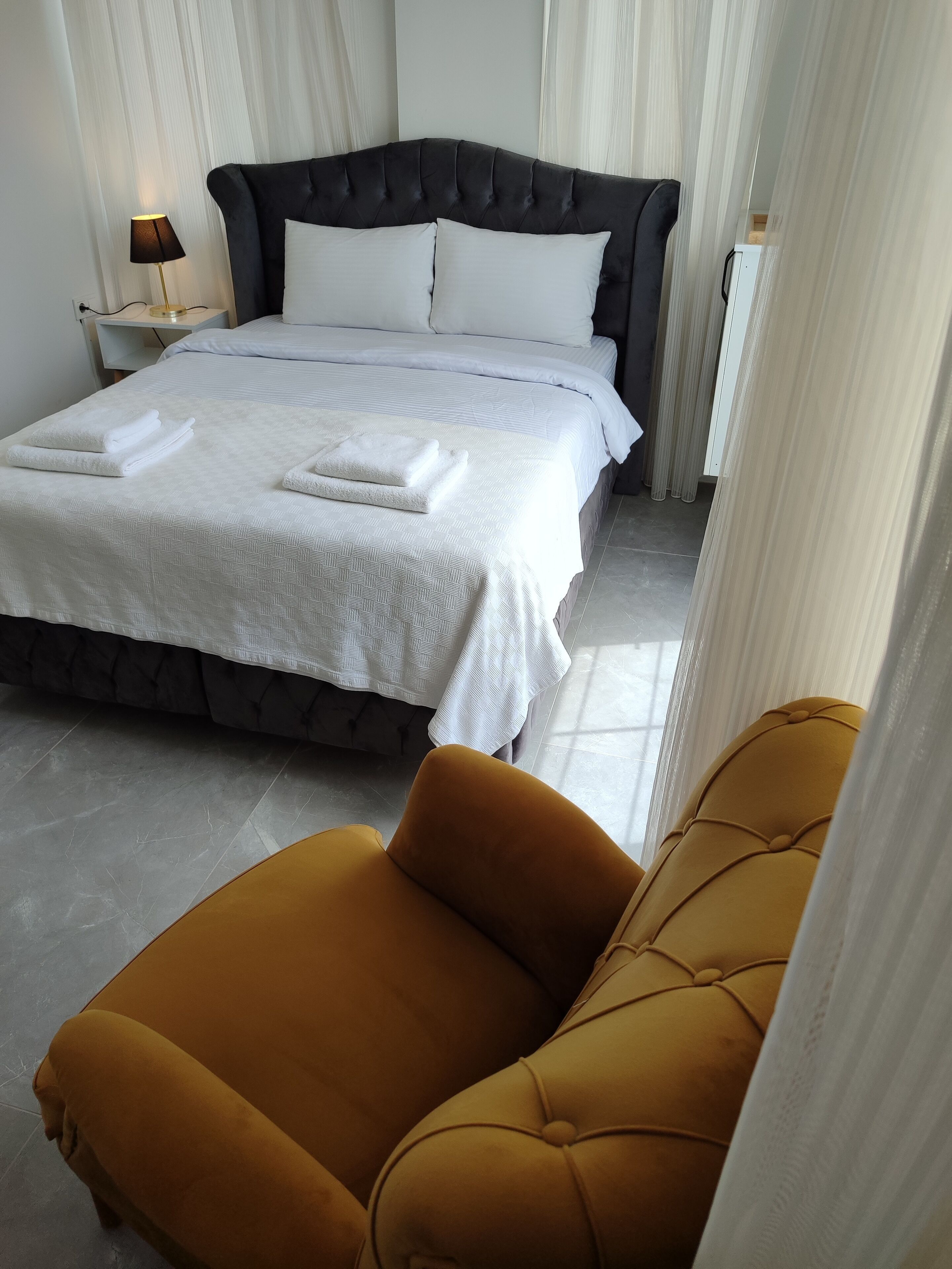Cama bem arrumada com lençóis brancos e toalhas dobradas, uma poltrona cor de mostarda e um abajur.