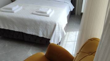 Cama bem arrumada com lençóis brancos e toalhas dobradas, uma poltrona cor de mostarda e um abajur.