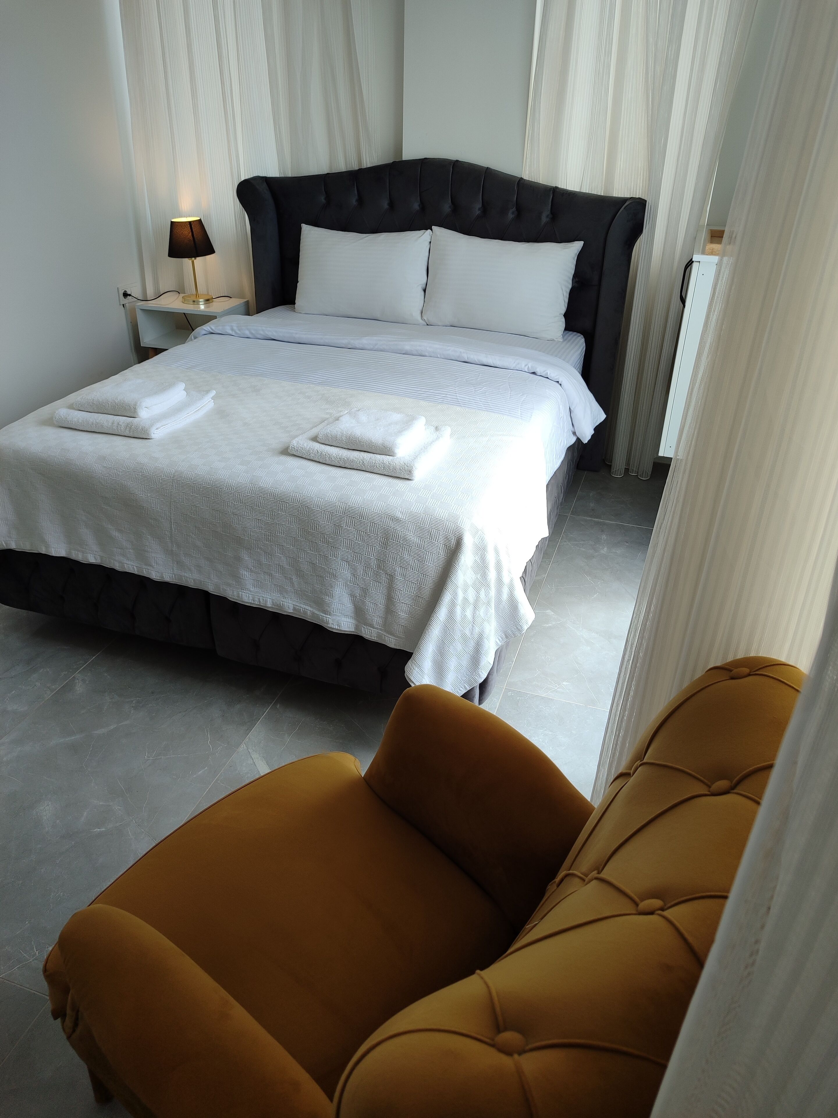 Una cama bien tendida con sábanas y toallas blancas, un cabecero tapizado en negro y un sillón de color mostaza en una habitación de hotel moderna.