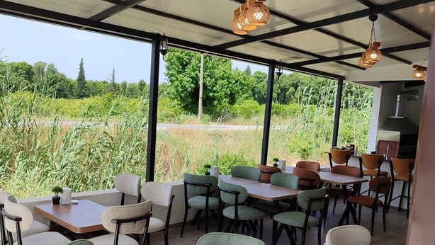 A covered outdoor seating area with tables and chairs, a round table with a glass top, and a view of a field through large windows. - The Dalaman Suites & Pension Dalaman Airport 10 km (Dalaman)