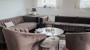 A living room with a sectional sofa, a round coffee table, and two armchairs. There is a window with curtains, a radiator, and a lamp on a side table. - Villa Jonny am Deich (Dornum)