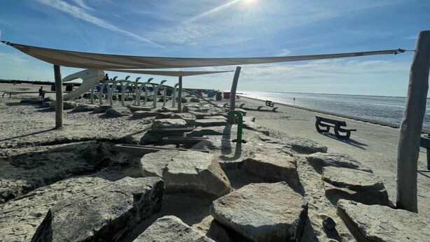 A beach with a shaded area, rocks, and a bench. - Villa Jonny am Deich (Dornum)