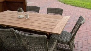 An outdoor patio with a wooden table and wicker chairs, overlooking a green field. - Villa Jonny am Deich (Dornum)